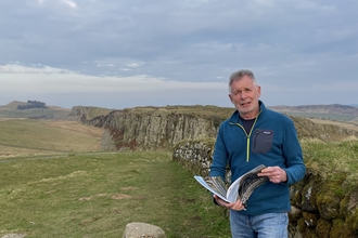 Image of author Ian Jackson taken from Steel Rigg with his book 'Northumberland Rocks' with Peel Cragg (part of the Whin Sill) in the background.