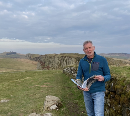 Image of author Ian Jackson taken from Steel Rigg with his book 'Northumberland Rocks' with Peel Cragg (part of the Whin Sill) in the background.