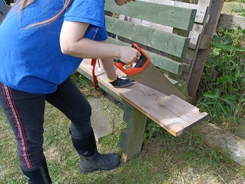 Raven is seen to be holding a saw in her right hand as it is placed onto the wood ready for sawing action.  