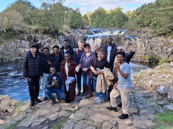 A group of people stand on rocky ground in front of a wide waterfall, with trees and flowing water in the background.