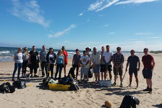 A group of volunteers standing together on a sandy beach, smiling and holding cleanup tools such as litter pickers and bin bags. Behind them, waves crash onto the beach under a clear blue sky.