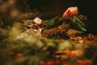 Close-up of hands crafting a rustic wreath with evergreen branches, red berries, pinecones, and natural decorations on a warmly lit worktable.