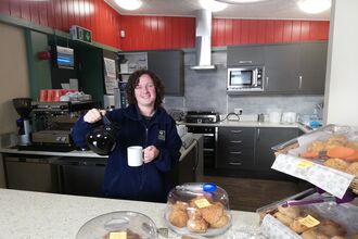 Person standing behind a café counter pours coffee into a mug, with covered cakes and pastries displayed in the foreground and a kitchen area in the background.