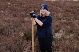 Blonde-haired woman standing on a nature reserve, wearing a blue beanie and work gloves.