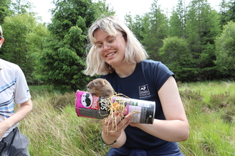 Image of Sally Johnson holding a watervole, which is standing on a recycled crisp tube, at Kielder forest.