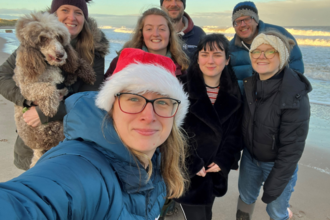 A group selfie photo of staff on a beach. A woman is in the foreground taking the selfie, wearing a Santa hat. Behind her is six other members of staff, one of which is carrying a dog. Everyone is wearing winter clothes and smiling.