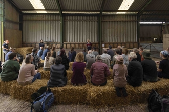 A large group of people sit on three rows of hay bales in a barn listening to a talk for Great North Bog Day 2024
