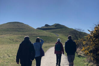 Four people walking away from camera towards Northumberlandia with blue skies above.