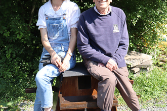 A man and woman sitting outside on a barn owl box in front of bushes.