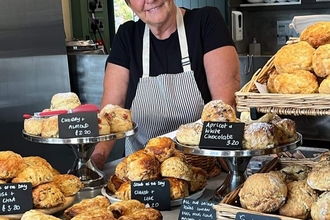 The picture shows a smiling woman standing behind a counter laden with scones and sausage rolls. She is wearing a black t-shirt and a striped apron. The food is displayed on tiered stands and in baskets, with small signs indicating their flavours and prices.