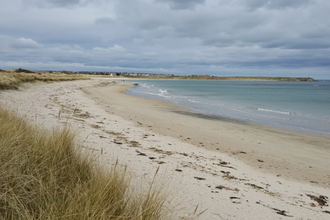Coastal landscape of a sandy beach, with dunes to the left of the image and the ocean to the right of the image. The sky is filled with heavy grey clouds.