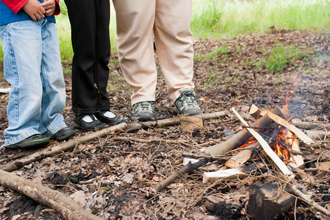 The image shows the lower portions of three people standing around a small campfire. 