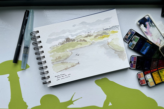 The image shows a sketchbook lying open with a watercolor painting of a landscape, possibly a lake or river scene with greenery and a few birds. Beside the sketchbook are a pen, a water brush, and a palette of watercolor paints. In the background, there's a green silhouette of a snail and a frog on a white surface. The handwriting in the sketchbook indicates the painting is of "Hauxley Discovery Centre."