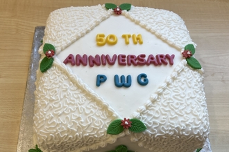 A square cake is seen on a silver stand; the cake is decorated with a white frosting design on the sides, with a border resembling lace. In the center, "50TH ANNIVERSARY" is written in red and yellow, with "PWC" below in blue icing. The cake is further decorated with red flowers and green leaves.
