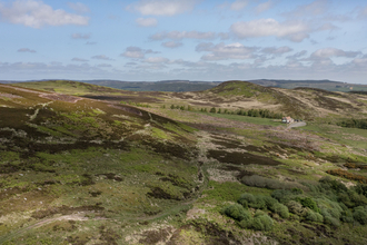 The image shows an expansive view of a rolling, grassy landscape under a sky dotted with clouds. The hills are covered with varied vegetation, including patches of green and areas that appear brownish-tan, possibly indicating different types of ground cover or different stages of growth.
