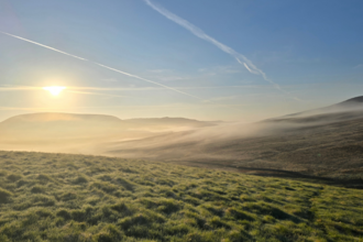 Vast green, grassy landscape with rolling hills. Fog is settled on the ground in the distance and the sun is rising above one of the hills to the left of the image. The sky is clear apart from two airplane trails.