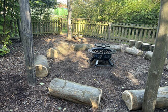Outdoor fire pit area with log seating arranged in a circle, surrounded by a wooden fence and trees.