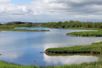 Freshwater lake with small grassy islands, surrounded by green vegetation under a cloudy sky.