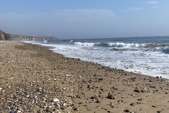 Pebble-covered sandy beach with gentle waves and distant cliffs under a clear blue sky.