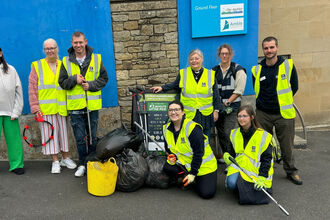 A group of people wearing high‑visibility vests pose together outdoors after a community litter pick, standing beside bags of collected rubbish and holding litter‑picking tools. A blue building wall and signage are in the background.
