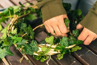Hands weaving green ivy into a circular wreath on a wooden surface.