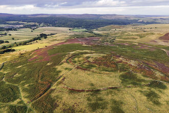 Aerial view of rolling countryside with patches of green, brown, and purple heathland, showing circular earthwork patterns on a hillside.