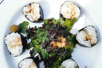 A white plate holds a circular arrangement of six sushi-style rolls made with seaweed and white rice, each filled with small pieces of vegetables or other ingredients. In the center of the plate is a mound of mixed green and dark seaweed, topped with additional finely shredded seaweed. The dish is presented simply on a light-coloured surface.