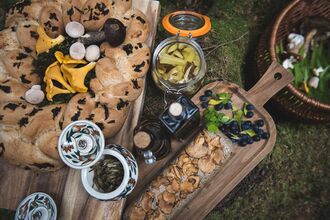 A rustic outdoor food spread displayed on wooden boards. There is a round loaf of bread with dark herbs baked into the crust, surrounded by assorted wild mushrooms. Nearby are jars of preserved vegetables, bottles of dark liquid (possibly oils or syrups), a long slice of bread topped with cooked mushrooms, and small piles of fresh berries and greens. Everything is arranged on a mossy forest surface.
