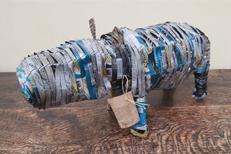 A small sculpture shaped like a hippopotamus, made from tightly layered and woven strips of recycled metal cans, displayed on a wooden surface.
