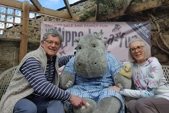 Two adults sit on a bench beside a large plush hippo mascot holding a small toy duck, with a charity event banner behind them in an outdoor garden setting.