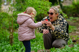 A young child in a pink jacket hands a small red container to an adult wearing sunglasses and a patterned jacket, both standing in a lush green garden.