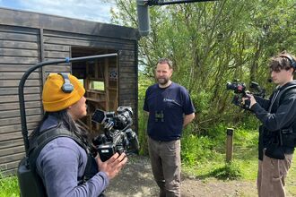 Film crew recording an outdoor interview beside a wooden wildlife hide, with a person speaking while two camera operators and a boom microphone capture audio and video.