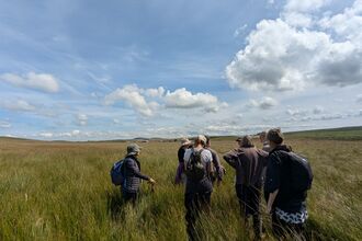 Group of people with backpacks walking through tall grass in an open countryside landscape under a wide blue sky with scattered clouds.