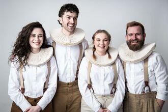 Four people standing side by side in a studio, wearing white shirts, suspenders, and large ruffled collars, with a plain light background.