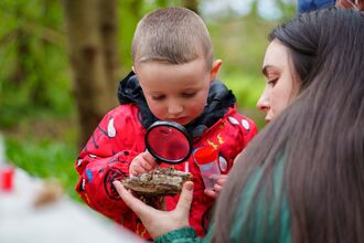 Child in a red jacket uses a magnifying glass to examine a piece of wood while an adult holds it, during an outdoor nature activity.