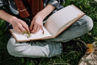 Hands placing small plants or seeds into an open blank notebook while sitting on grass outdoors, with garden items nearby.