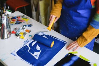 Person using a brayer to roll blue ink onto a printmaking plate on a cluttered table with paint tubes and brushes.