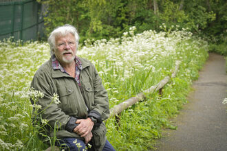 Bill Oddie OBE sitting next to long grass. Image by Chris Taylor Photography.