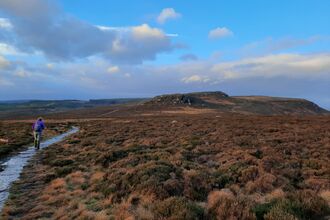 A person with a backpack walks along a stone path through open moorland toward a rocky hill beneath a wide blue sky with large clouds.