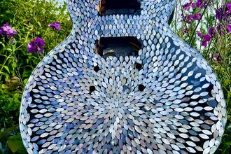 Decorative electric guitar covered in shimmering shell mosaic, standing among purple flowers and green garden foliage under a blue sky.