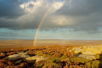 Bright rainbow stretching across a rocky moorland under dramatic dark clouds, with sunlight illuminating the landscape and a faint second rainbow visible in the distance.