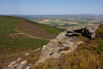 Panoramic view from a rocky hillside overlooking patchwork fields, rolling hills, and forested slopes under a clear blue sky.