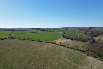 Aerial view of open countryside with green fields, hedgerows, scattered trees, and a small farmstead under a clear blue sky.