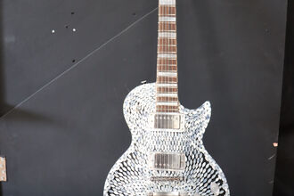 Decorative electric guitar with a iridescent pearl ovals radiating from the centre, standing on a floor stand against a dark wall.