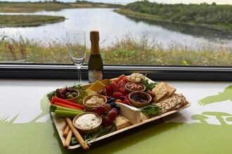Charcuterie board with cheese, fruit, crackers and a bottle of prosecco on a windowsill overlooking a tranquil lake and greenery.
