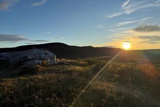 Sun setting over rolling hills, with rocky grassland in the foreground and wispy clouds in a clear blue sky, casting warm golden light across the landscape.