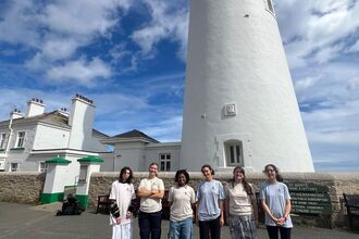Six young people standing in a line in front of a tall, white lighthouse. Behind the lighthouse is a blue sky with lots of white, fluffy clouds. 