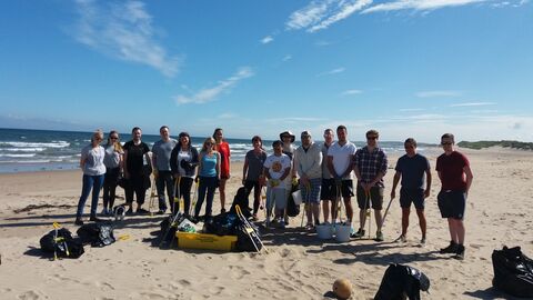 A group of volunteers standing together on a sandy beach, smiling and holding cleanup tools such as litter pickers and bin bags. Behind them, waves crash onto the beach under a clear blue sky.