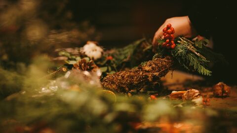 Close-up of hands crafting a rustic wreath with evergreen branches, red berries, pinecones, and natural decorations on a warmly lit worktable.