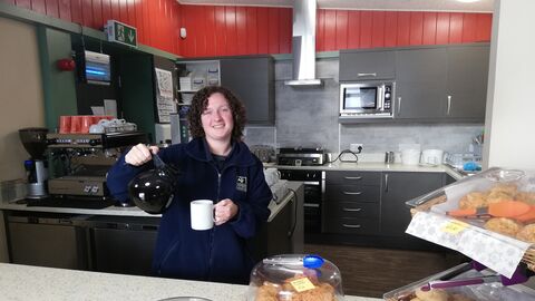 Person standing behind a café counter pours coffee into a mug, with covered cakes and pastries displayed in the foreground and a kitchen area in the background.